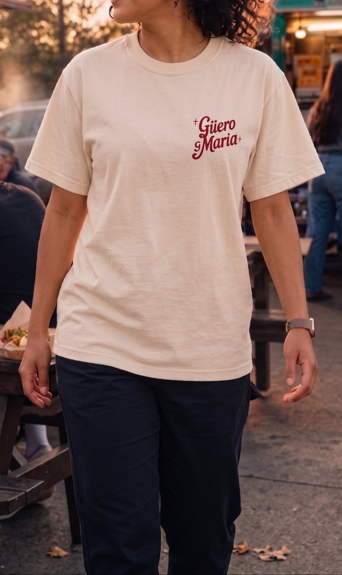 A woman wearing a cream-colored t-shirt with 'Gúero y Maria" printed in red cursive on the chest, standing in an outdoor street market setting.