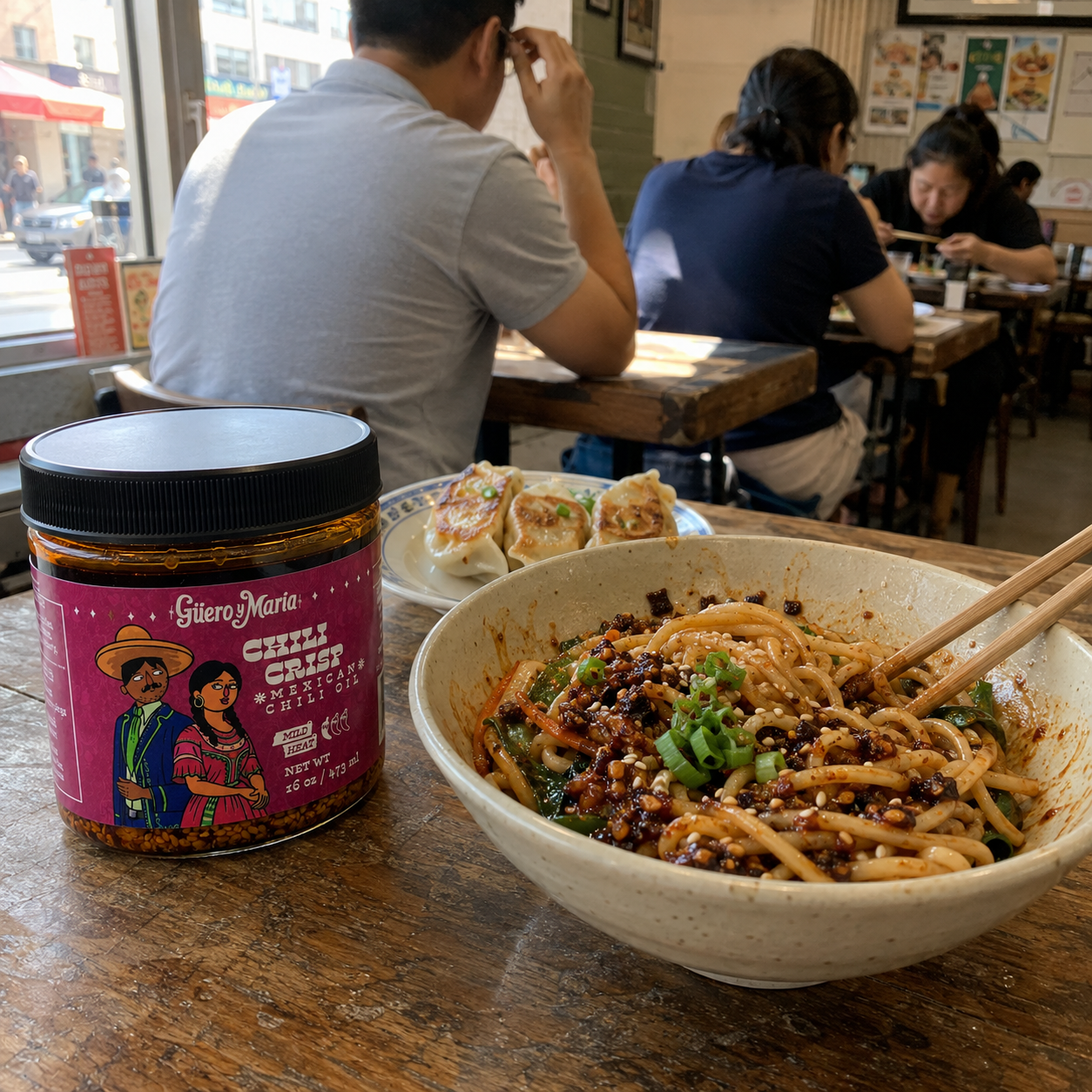 A jar of Giuero y Maria;s Chili Chip chilli oil  sits on a wooden table in a restaurant, next to a bowl of chili noodles with green onions and a plate of dumplings, with customers seated in the background.