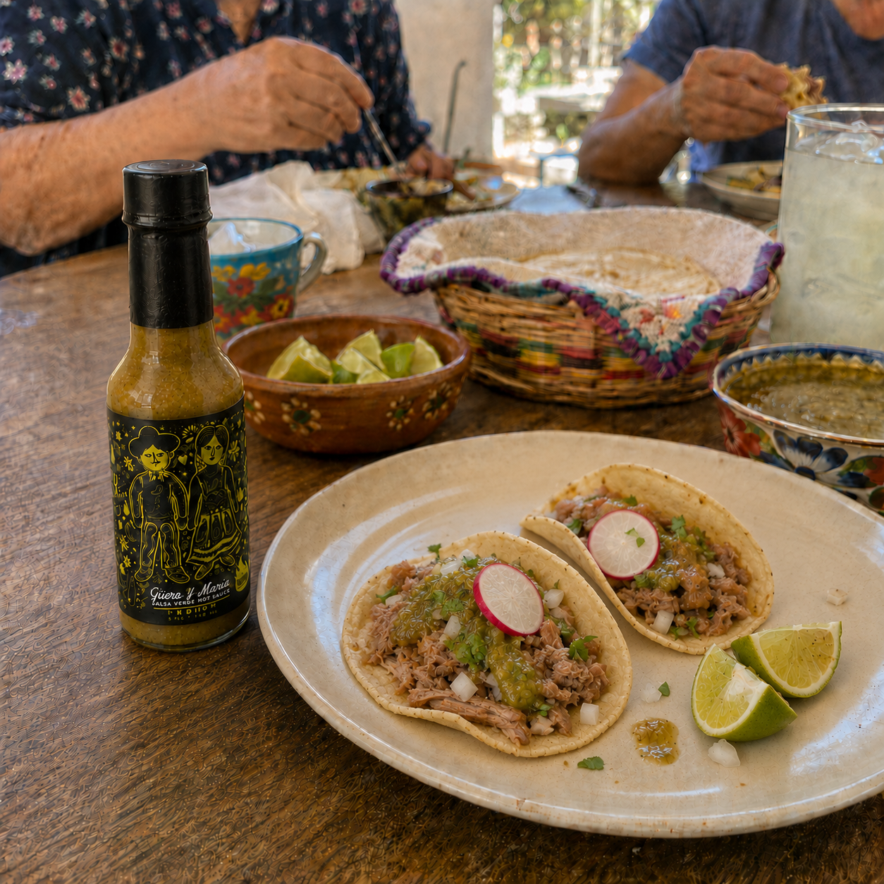 A bottle of Guero y Maria's salsa verde  hot sauce sits on a rustic wooden table next to a plate of two carnitas tacos with lime wedges and radishes, with people dining in the background.
